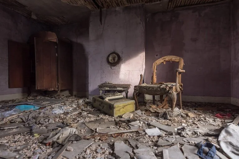 A dilapidated room filled with debris, broken furniture, and rubble on the floor; purple walls with a clock and a damaged ceiling.