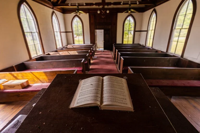 Open hymn book on a wooden pulpit in a small church with wooden pews and large arched windows.