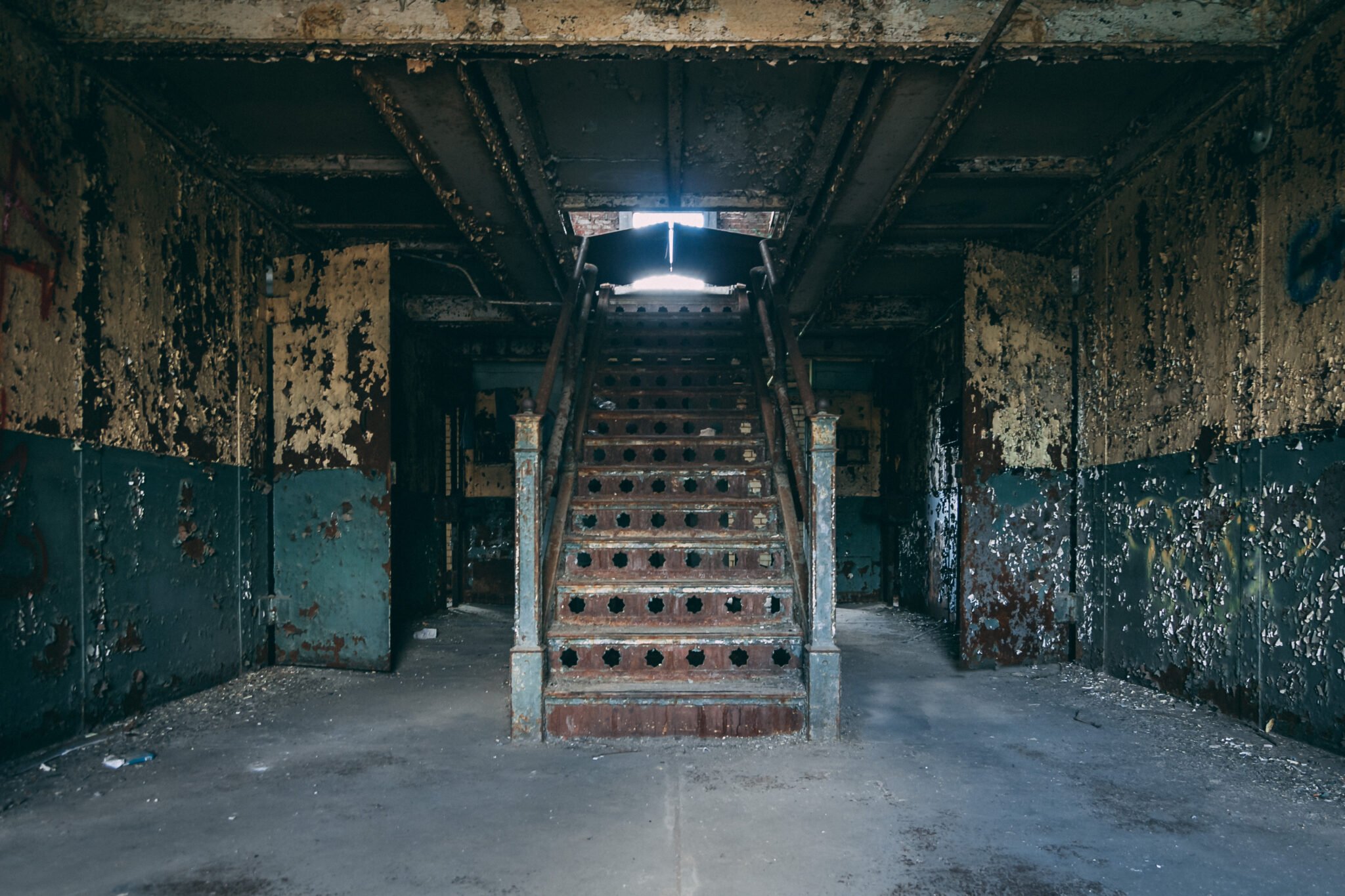 A set of old, rusted stairs inside a dark, abandoned building with peeling yellow and green walls and debris on the floor.