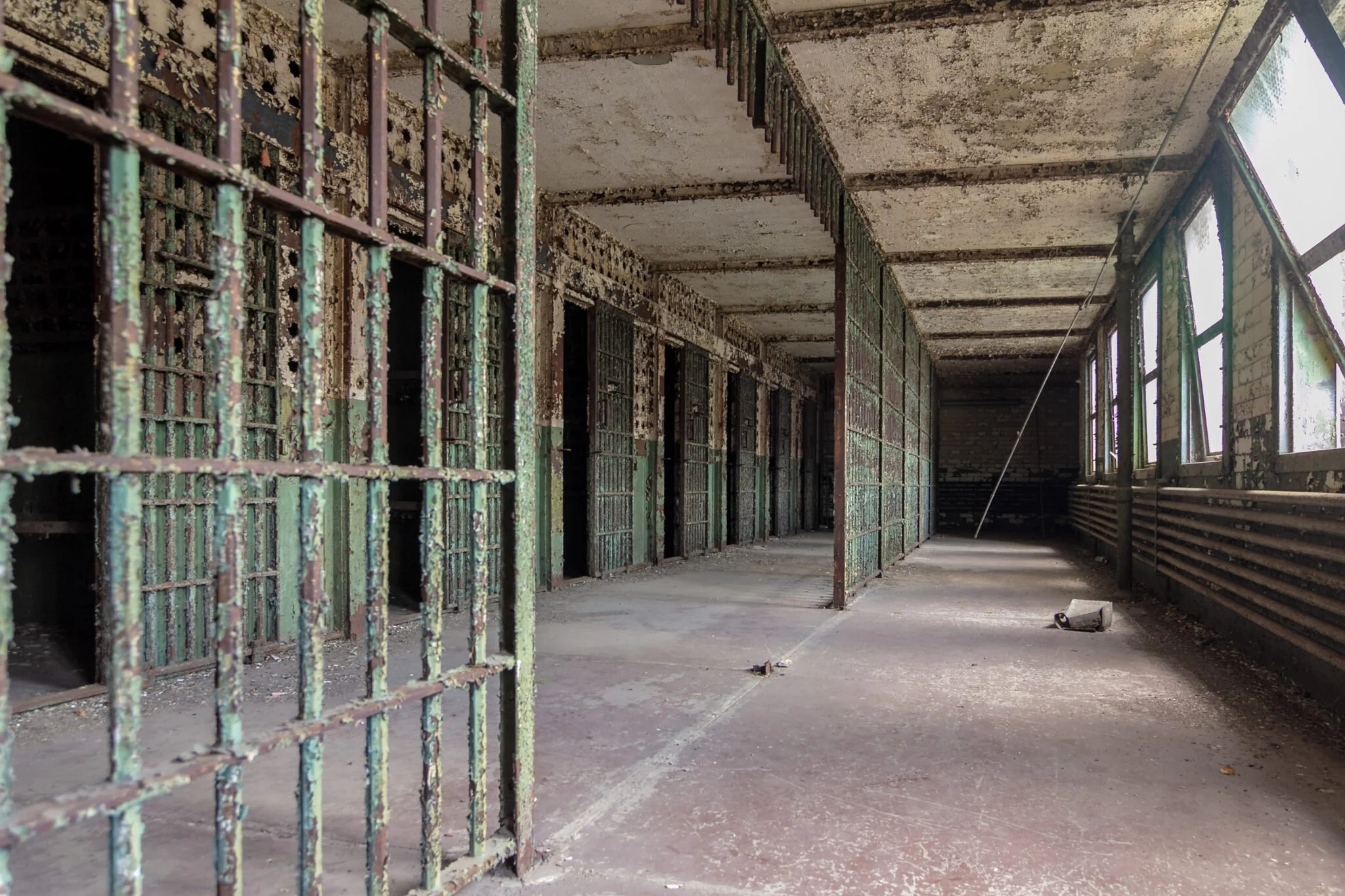An abandoned prison cellblock with rusted metal bars and peeling paint on walls and ceilings, showing signs of neglect and decay.