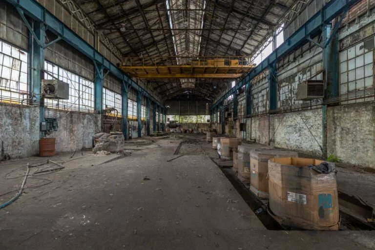 An empty, abandoned industrial warehouse with a high arched ceiling, large windows, and scattered debris and boxes on the floor.