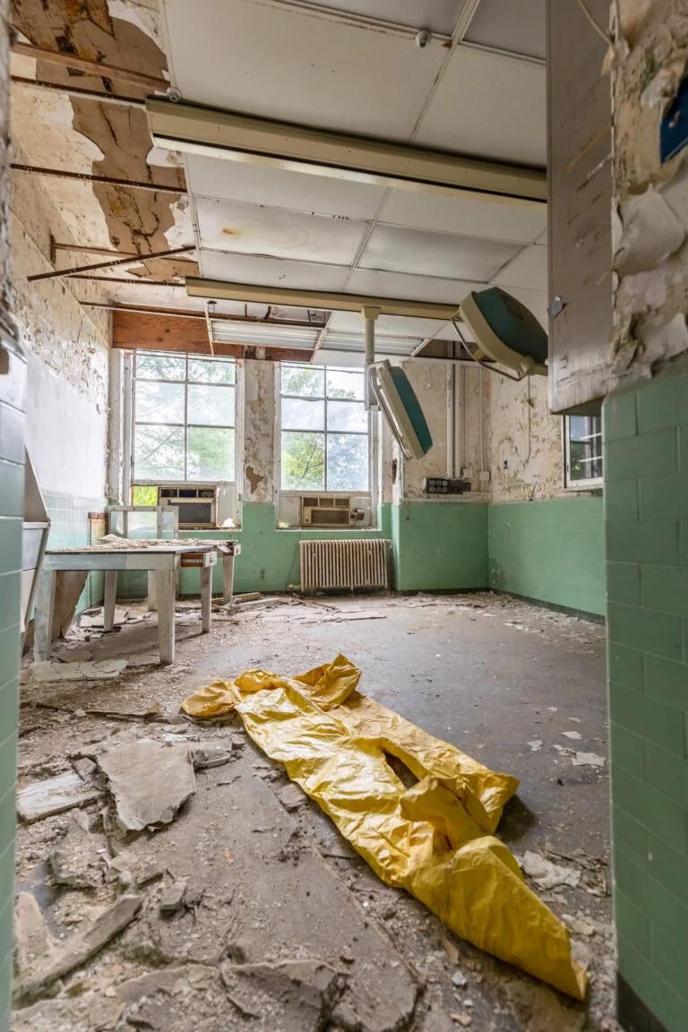 An abandoned room with peeling paint and debris on the floor, containing broken ceiling tiles, a yellow bag, and old furniture.