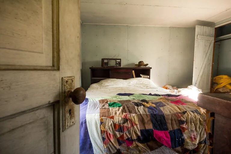 A rustic bedroom with an unmade bed covered by a colorful patchwork quilt. There is a small dark wood headboard with a vintage radio and a hat on top. The room has a wooden door slightly open revealing yellow cloth on the side, and the walls and ceil