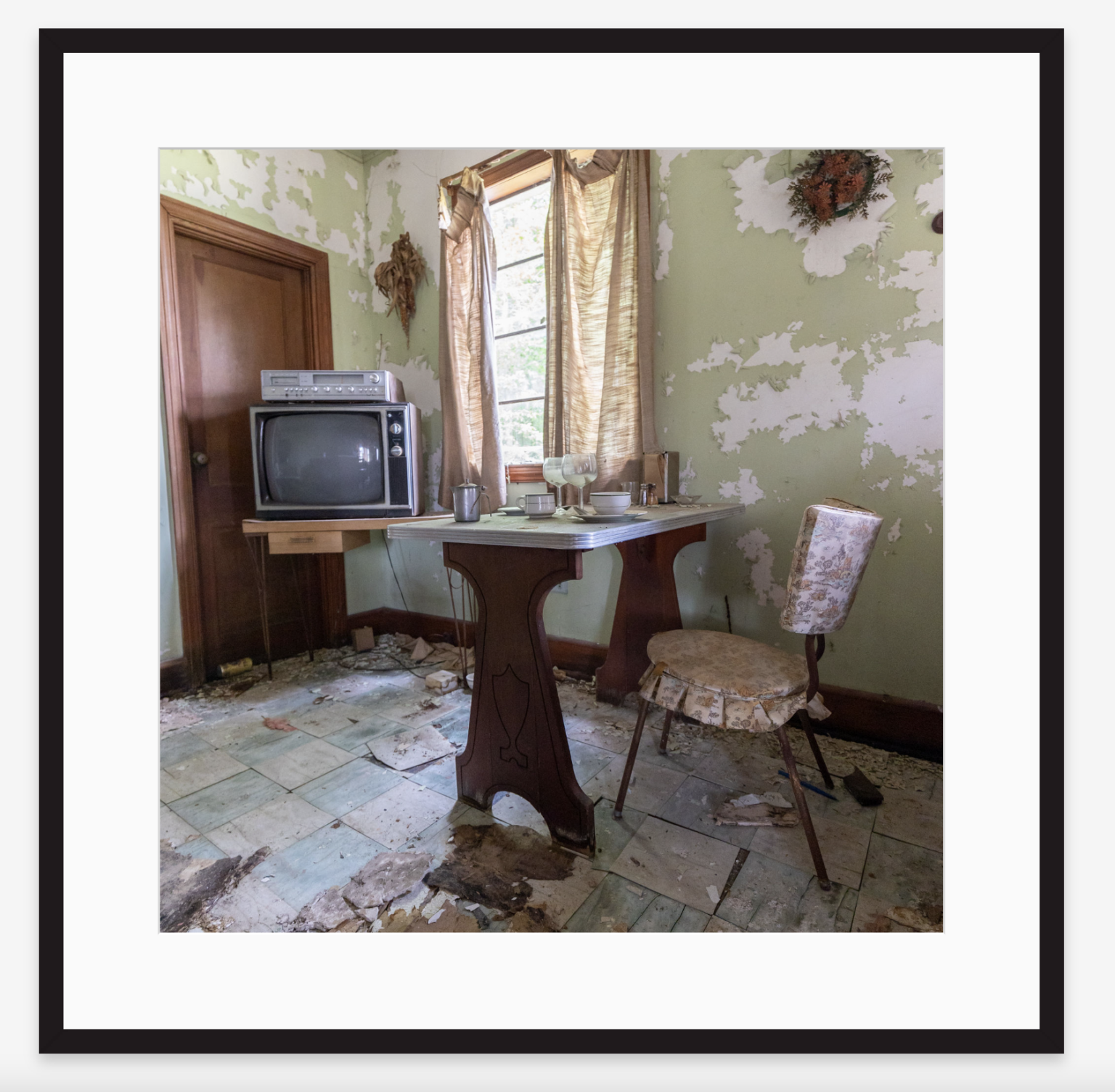 A room with peeling green paint on the walls, a small table with dishes, missing floor tiles, an old TV on a stand, a worn-out chair, and a window with torn curtains.