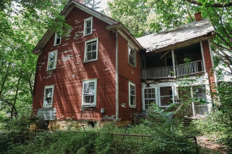 An old, abandoned two-story brick house surrounded by overgrown vegetation and trees.
