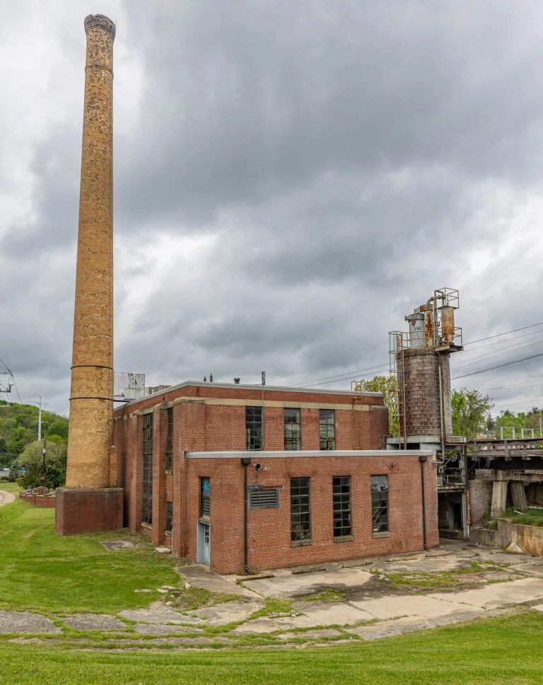 An old industrial building with a tall brick chimney, rusted machinery, and a large smokestack under a cloudy sky.
