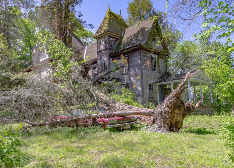 An old, abandoned Victorian-style house with a steep roof and turrets, surrounded by trees. A large uprooted tree is fallen in the front yard, damaging part of the house and a red vehicle underneath.
