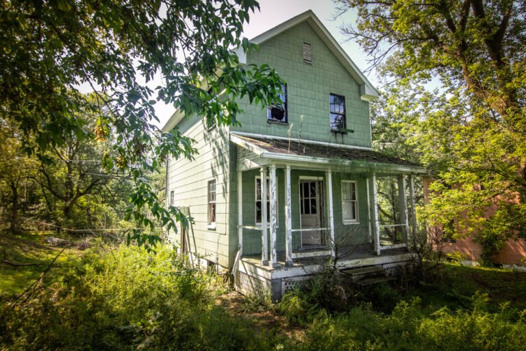 Old, abandoned green house with a porch, surrounded by overgrown trees and bushes.