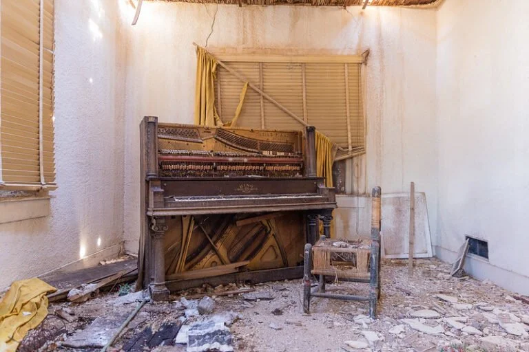 An abandoned, dusty room with a worn upright piano and a wooden chair with peeling paint and debris on the floor.