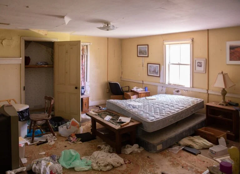 A messy bedroom with a stained carpet, a cluttered bed with a mattress and box spring, a wooden nightstand with a lamp, a small desk with a chair, a window with blinds, and various items scattered on the floor.