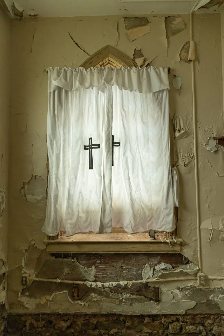Old, peeling interior wall with damaged drywall and peeling paint, covering a backstage or closet space with a small platform and a curtain with crosses covering an entryway.