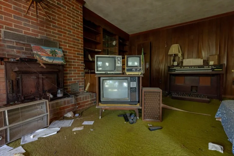 A living room with a brick fireplace, old TV sets stacked on top of each other, a vintage speaker, a piano with a lamp and objects on top, and scattered papers and remote controls on a green carpet.