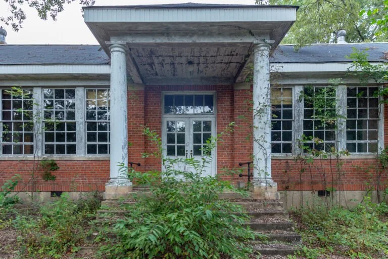 An old, abandoned brick house with large boarded-up windows and overgrown vegetation. The front porch has four columns and steps leading up to a door with glass panes.