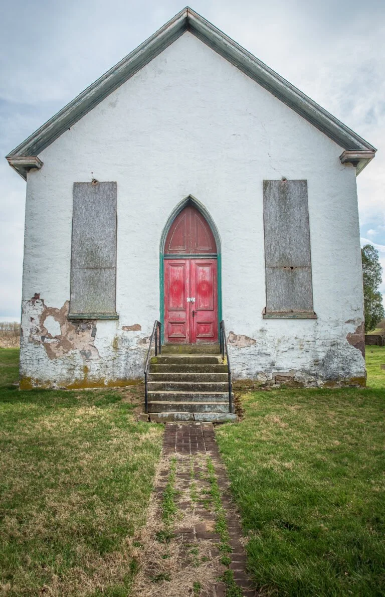 Old white church with a red arched door, boarded-up windows, and peeling paint, with steps leading up to the entrance and a brick pathway in front.