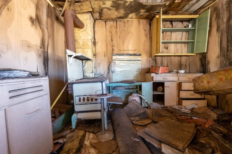 An old, abandoned kitchen with peeling wood walls, a rusted stove, and broken furniture covered in debris.