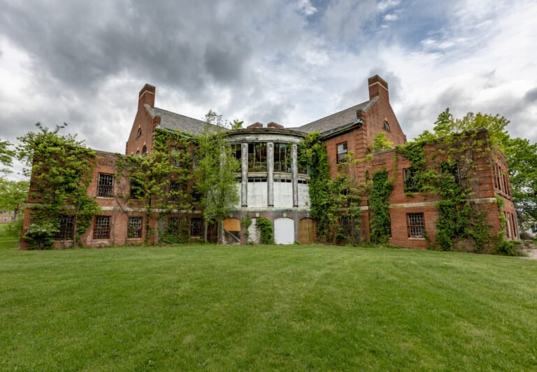 Abandoned brick mansion with overgrown vines and broken windows, situated on a grassy lawn under cloudy sky.