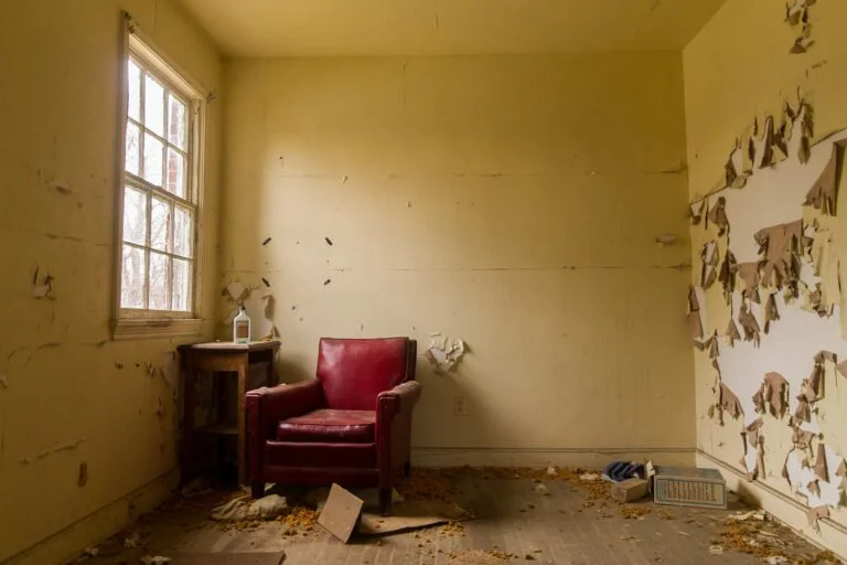 A small, empty room with peeling yellow walls, a single window on the left, and a red armchair in the corner. The floor is covered in debris, and the walls are damaged with peeling paint.