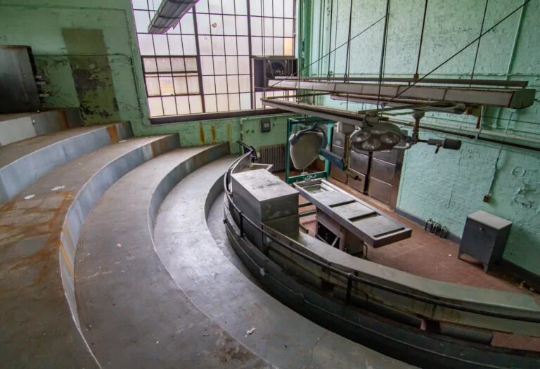 Empty old lecture hall with curved wooden seating, large windows, and worn painted walls.