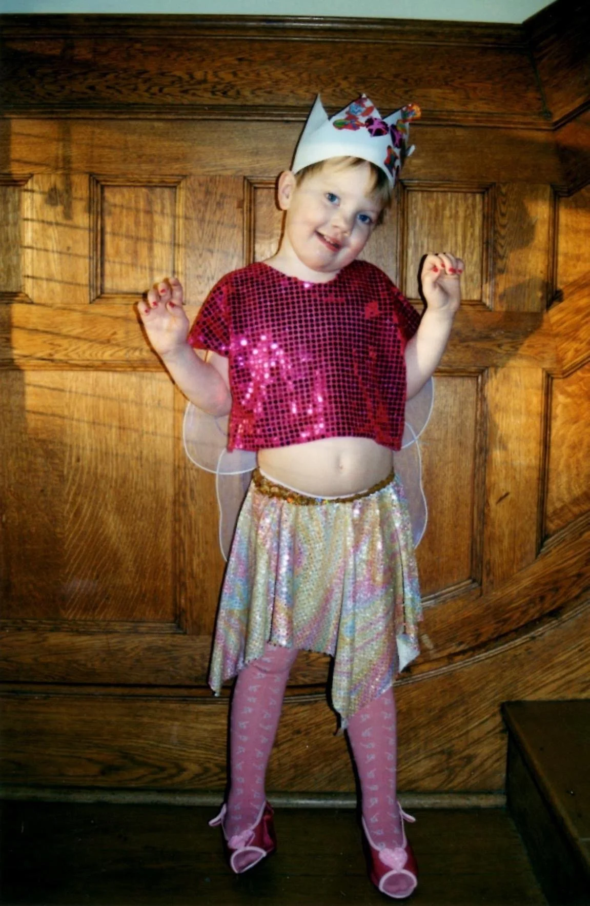 A young girl dressed in a fairy costume with pink sequined top, gold skirt, fairy wings, pink tights, and fairy shoes, wearing a colorful paper crown, standing indoor in front of wooden paneling.