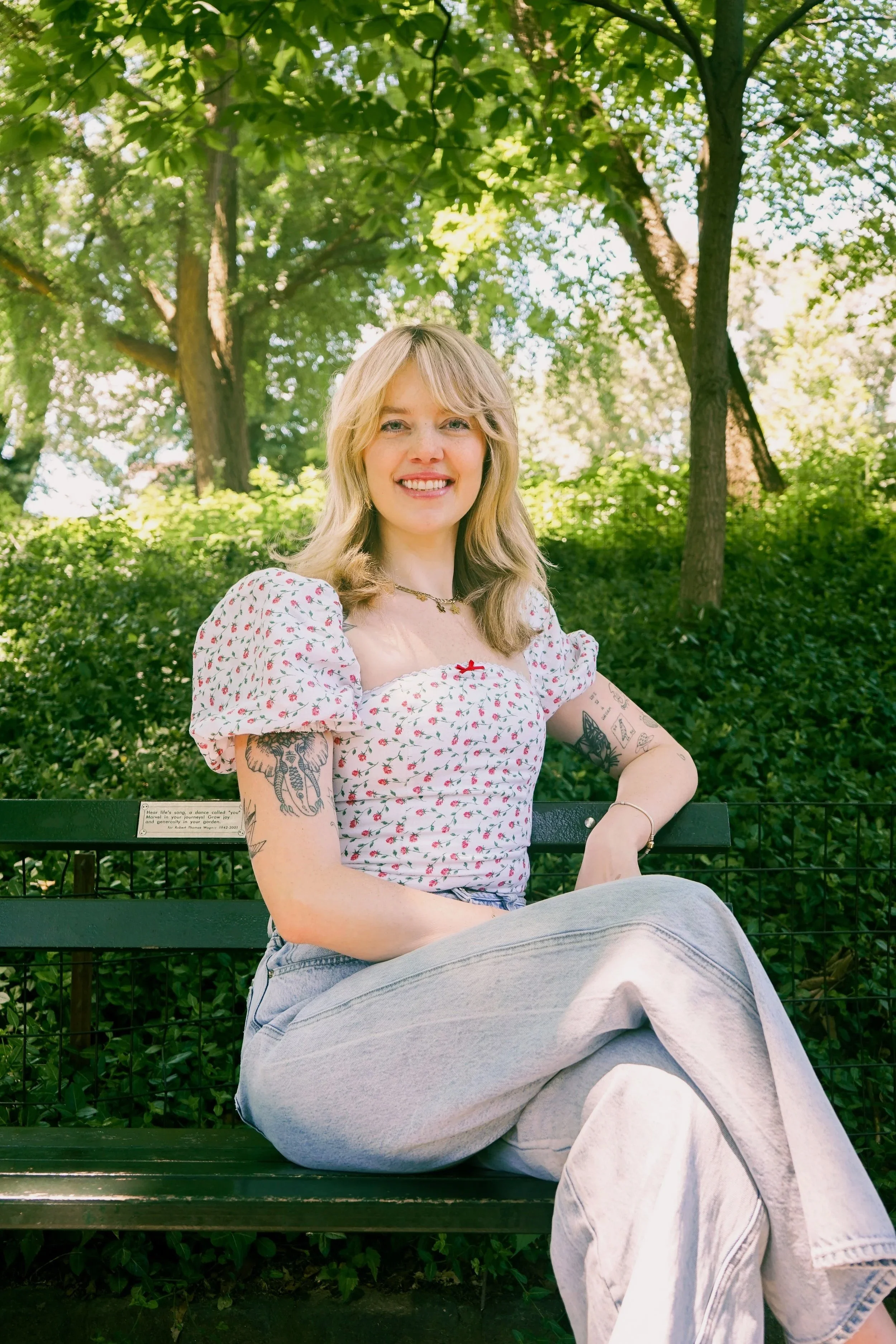 A young woman with blonde hair and tattoos on her arms sitting on a park bench. She is smiling and wearing a white, short-sleeved blouse with a red pattern and light-colored jeans. The background shows lush green trees and shrubbery in a park setting.