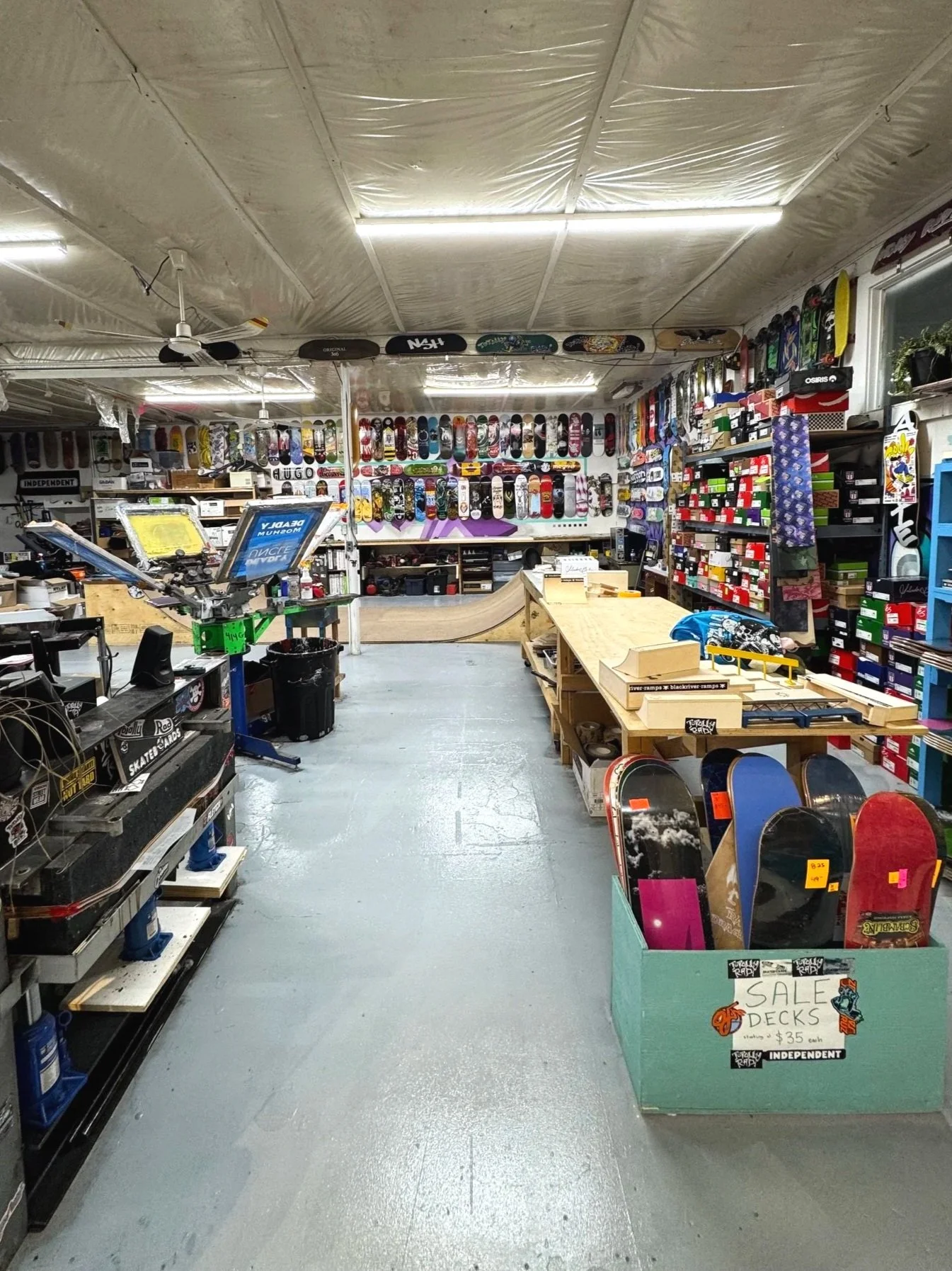 Interior of skate shop with skateboards on display on the walls and shelves, a checkout counter, and sale decks in a green cardboard box in the foreground.
