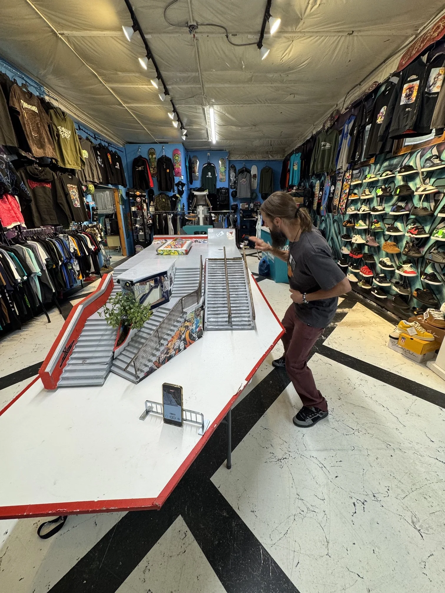 A man in a black shirt and maroon pants stands at a store checkout counter looking at his phone. The store has clothing and skateboards on the walls, shelves with sneakers on the right, and shirts on display on the left side. The ceiling is covered with beige fabric, and the floor has a black and white marble pattern.