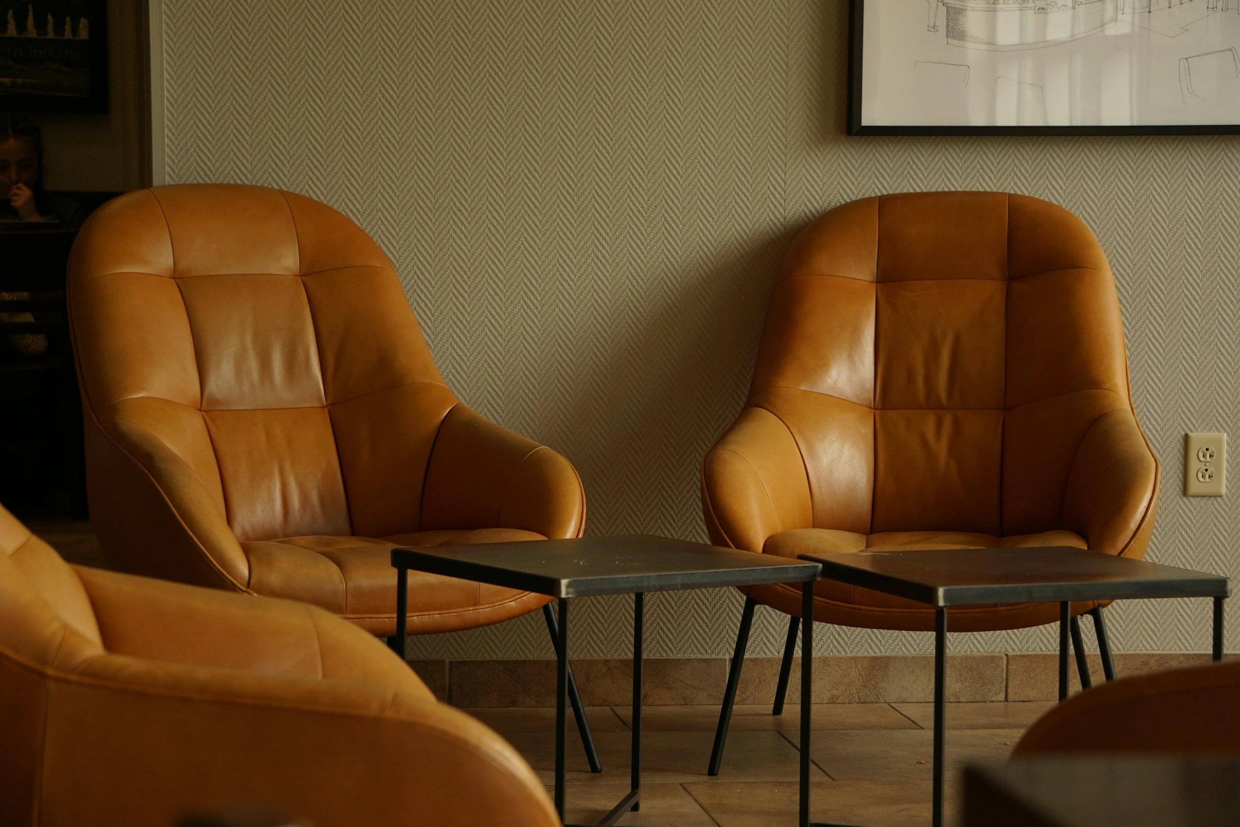 Two tan leather armchairs facing each other in a sitting area, with a black coffee table in between, against a patterned beige wall with framed artwork.