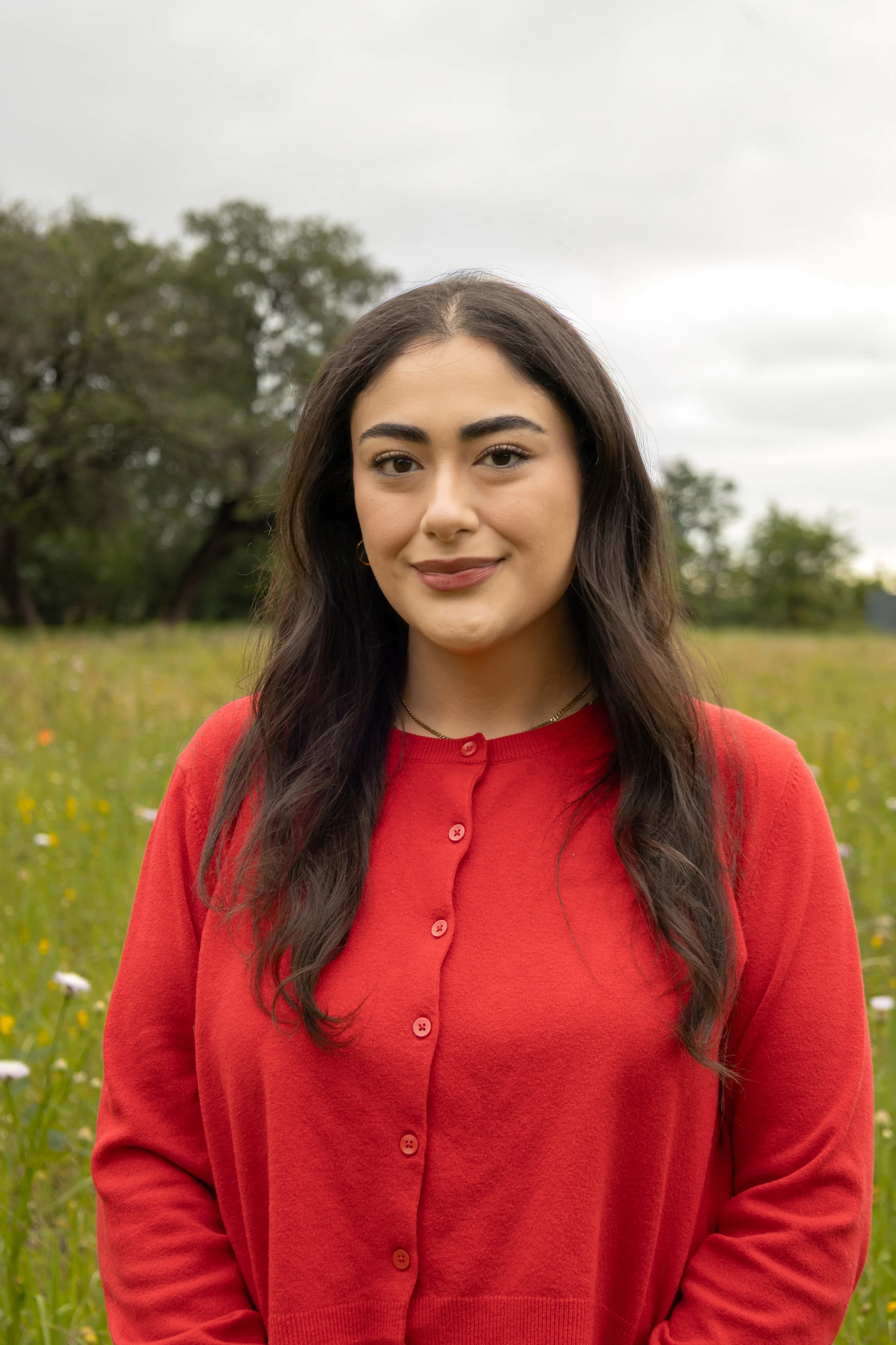A woman with long dark hair and a red sweater standing outdoors in a grassy area with trees in the background.