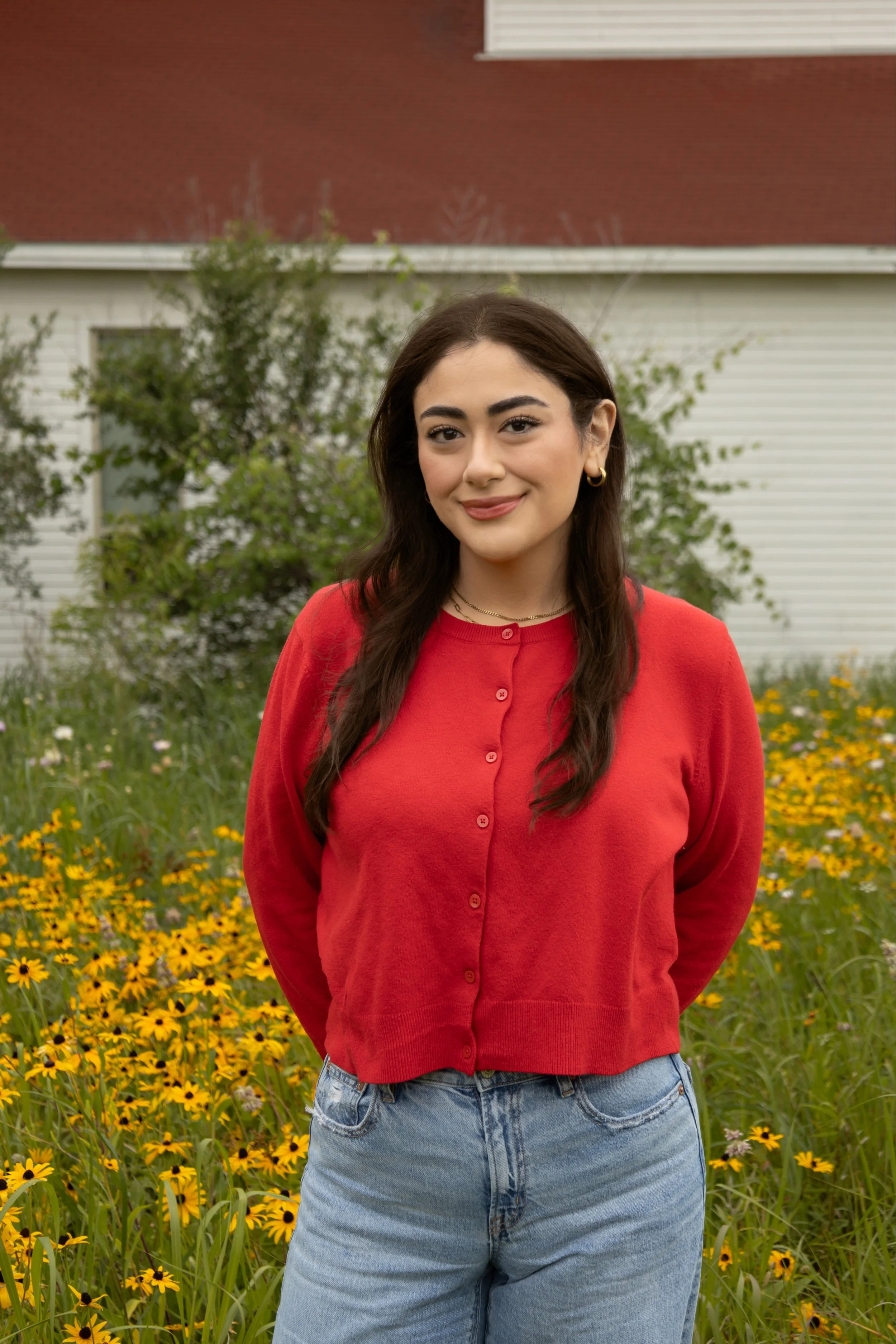 A young woman with long dark hair, wearing a red cardigan and blue jeans, standing outdoors in a field of yellow flowers with a red and white house in the background.
