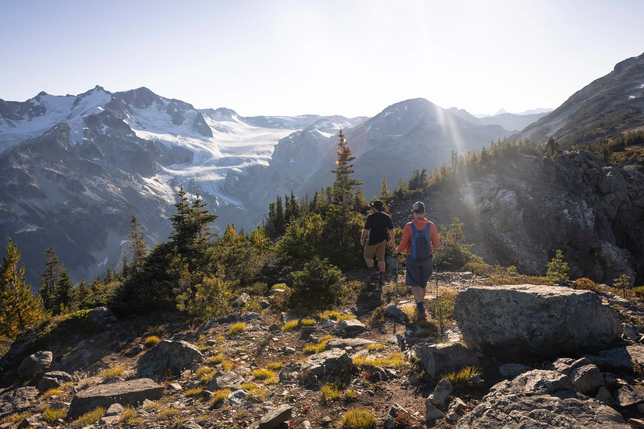 Two hikers walk away from us on a rocky trail, toward snow-capped mountains and bright sun. The picture is the next chapter: the step taken, the nerve found, moving toward a brighter stretch of the journey.