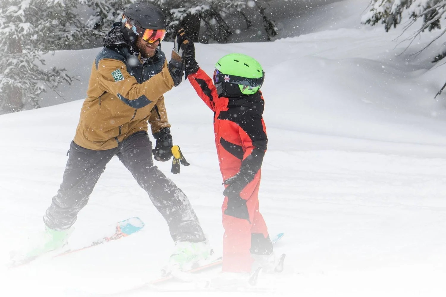 A parent and child high-five on skis in falling snow