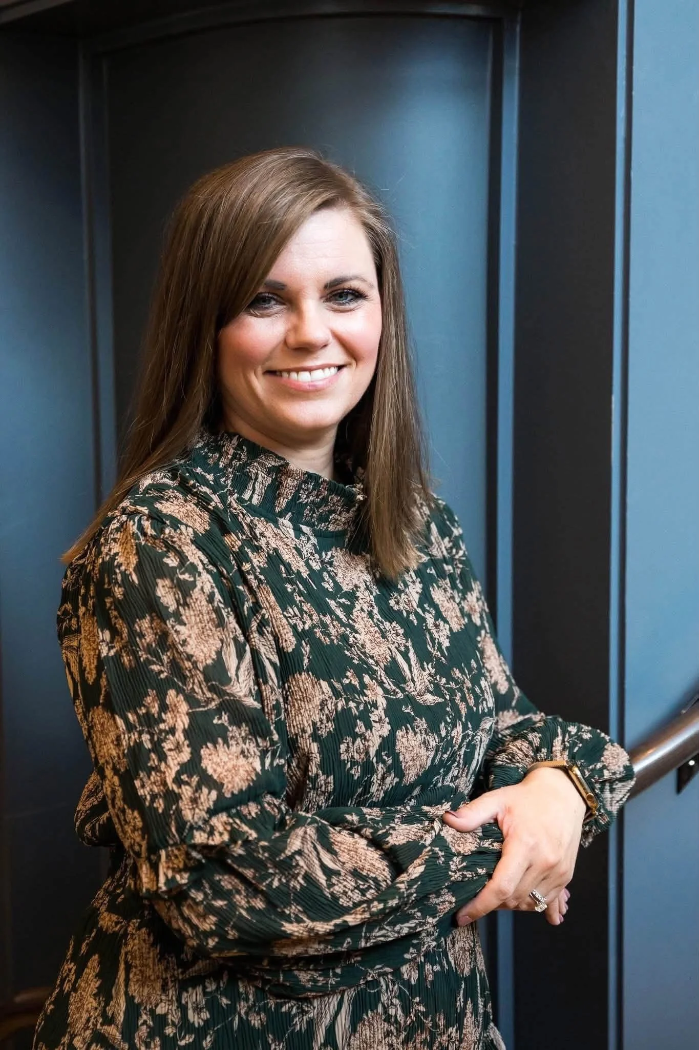 A woman with shoulder-length brown hair, wearing a long-sleeve dress with a floral pattern, smiling and standing near a blue elevator wall.