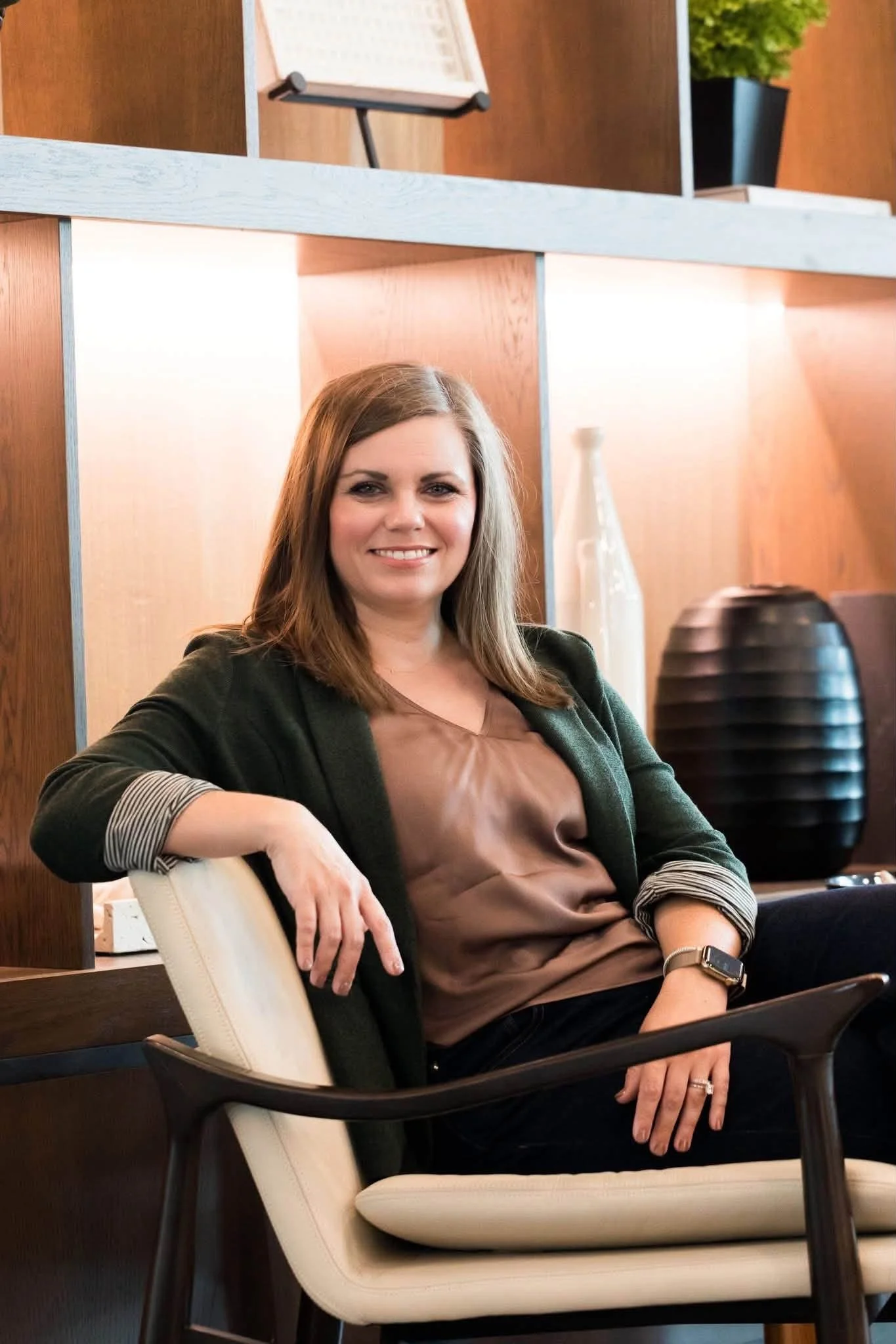 A woman with long brown hair, wearing a brown top and a dark blazer, sitting on a cream-colored chair with black arms in a modern office setting. She is smiling and sitting with one arm resting on the chair's armrest.