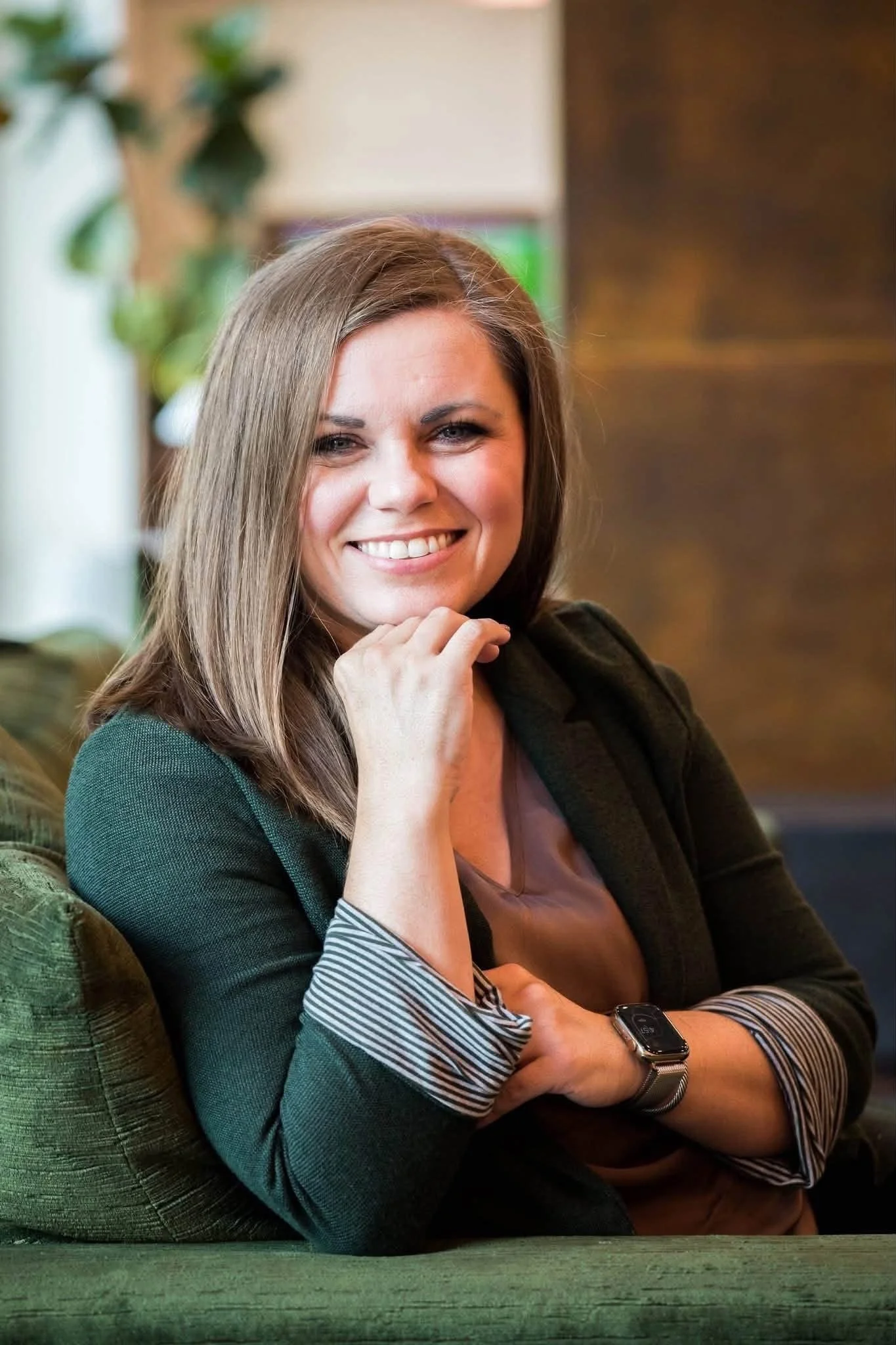 A smiling woman with long brown hair, wearing a green jacket and striped shirt cuffs, sitting at a table with her hand touching her face, in a cozy indoor setting with warm lighting.