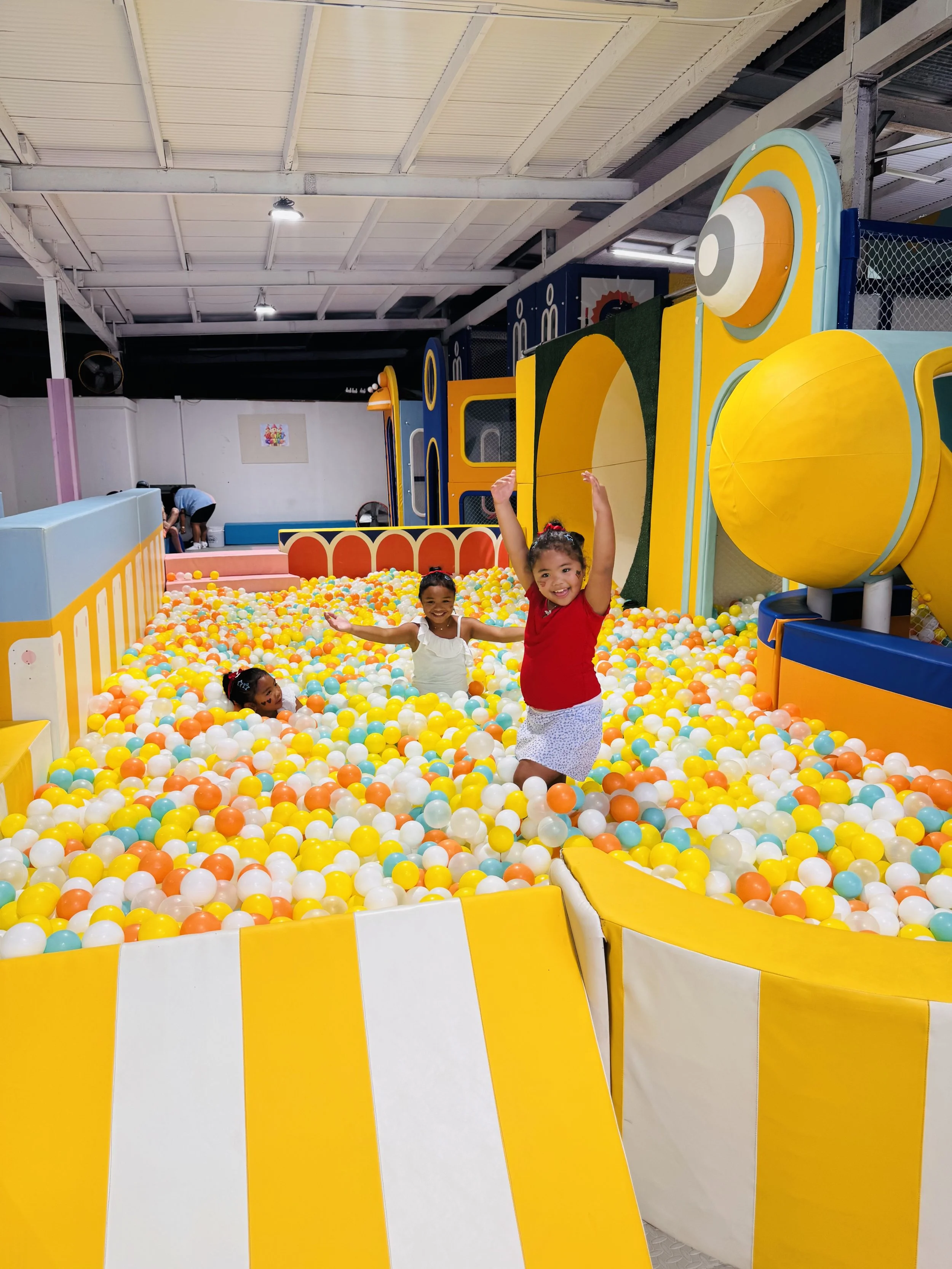 Children playing and having fun in an indoor ball pit with yellow, orange, white, and blue balls, featuring a large yellow and blue structure that resembles a train or vehicle.