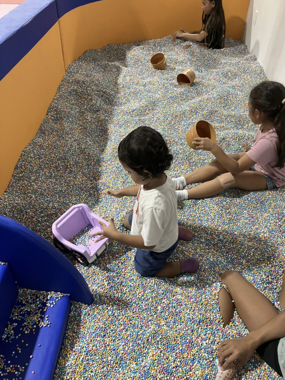 Children playing in a large indoor ball pit filled with multicolored small plastic balls, with some children sitting and others kneeling, and several orange buckets along the edge.