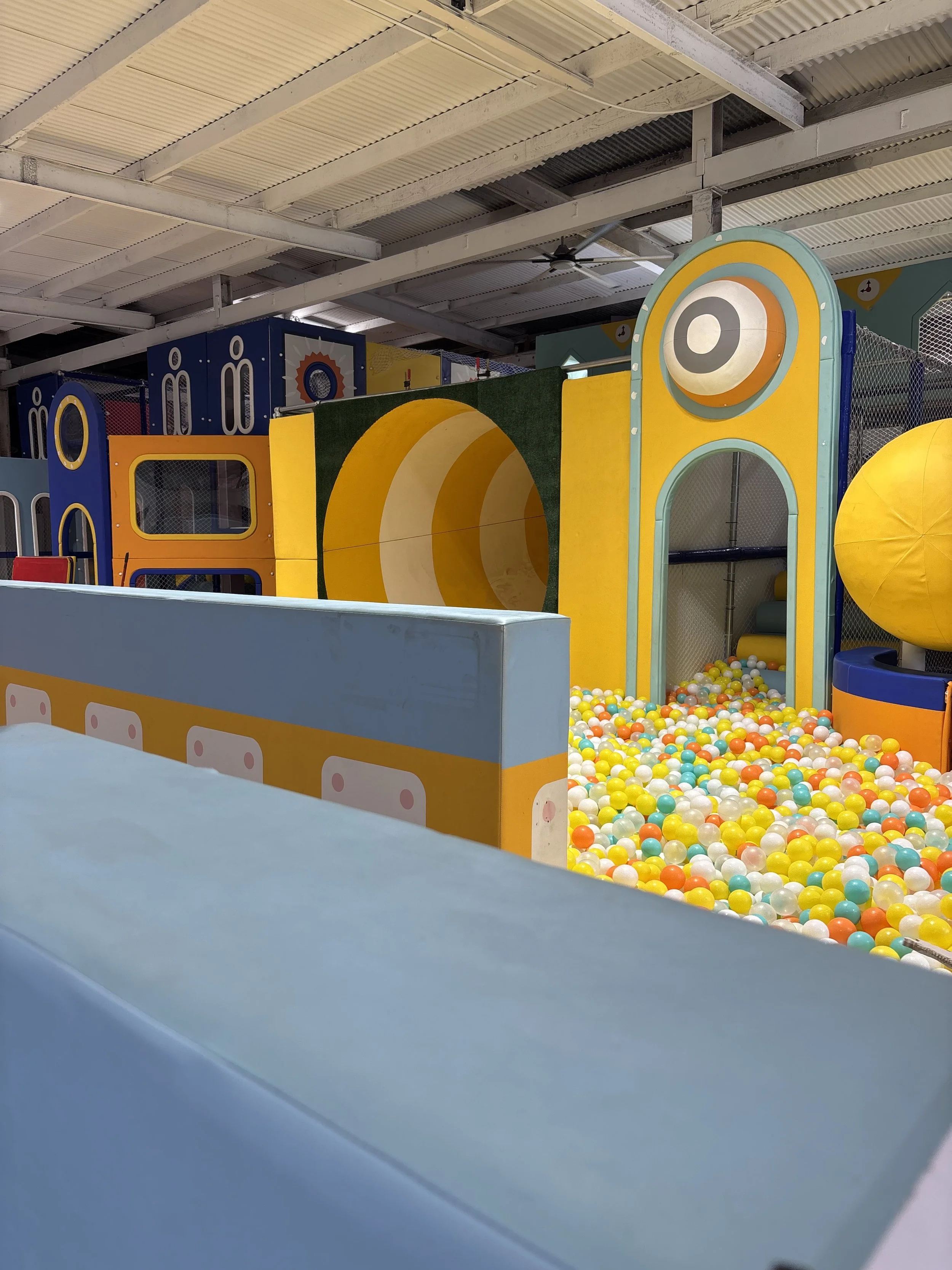 Indoor playground with colorful structures, including a ball pit filled with multicolored plastic balls, and various play zones with different colors and designs.