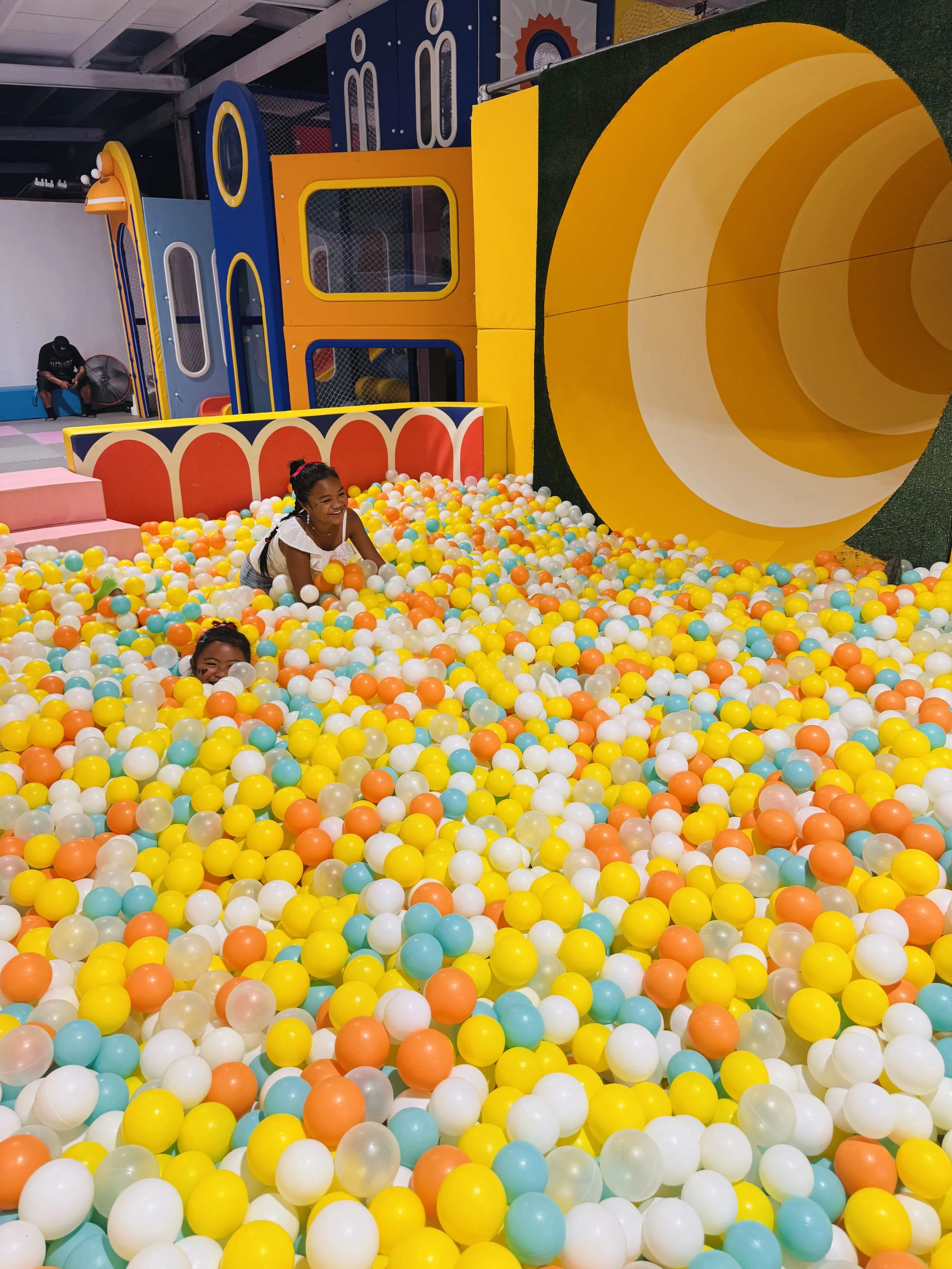 Children playing in an indoor ball pit with colorful plastic balls and a slide structure in the background.