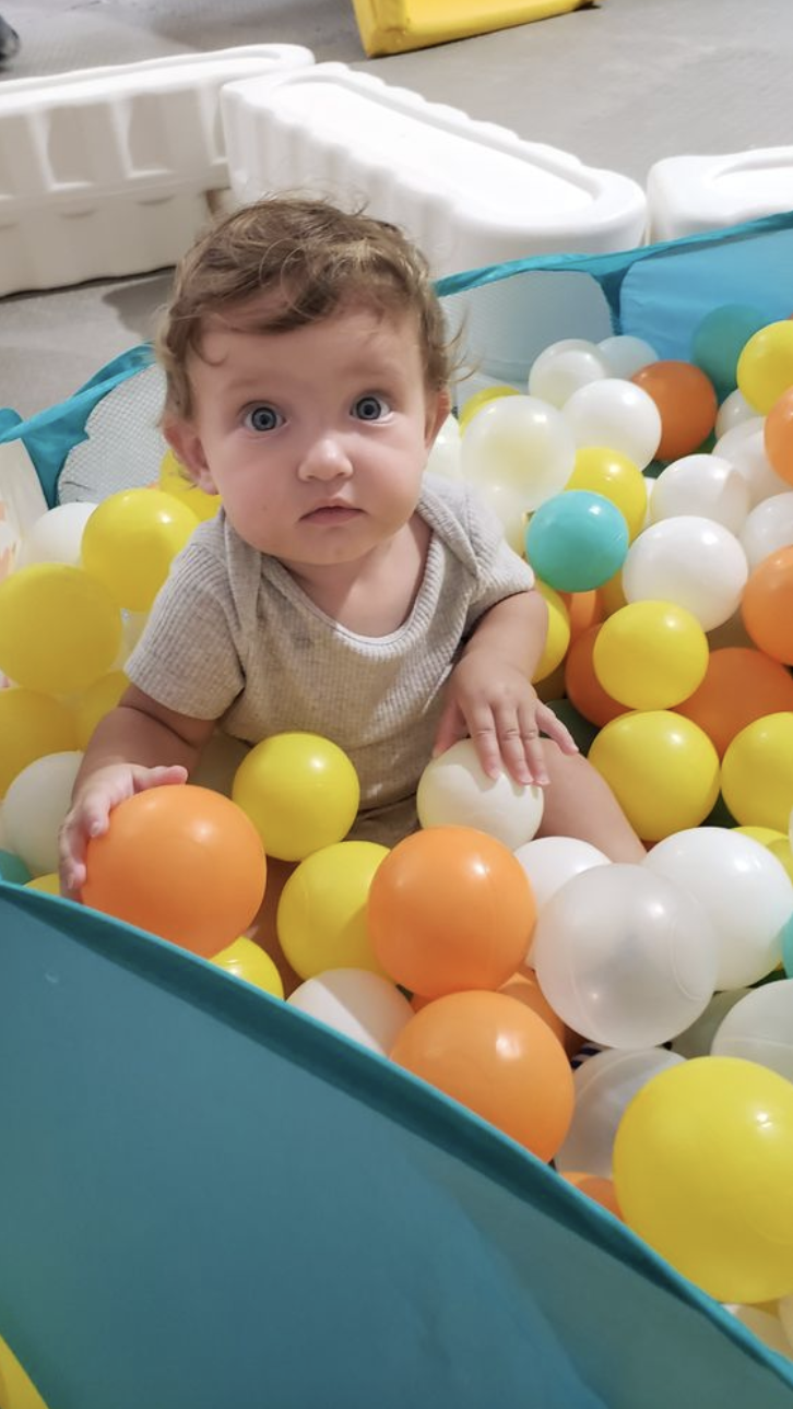 A young child with curly hair and blue eyes sitting in a ball pit filled with yellow, white, orange, and teal plastic balls.