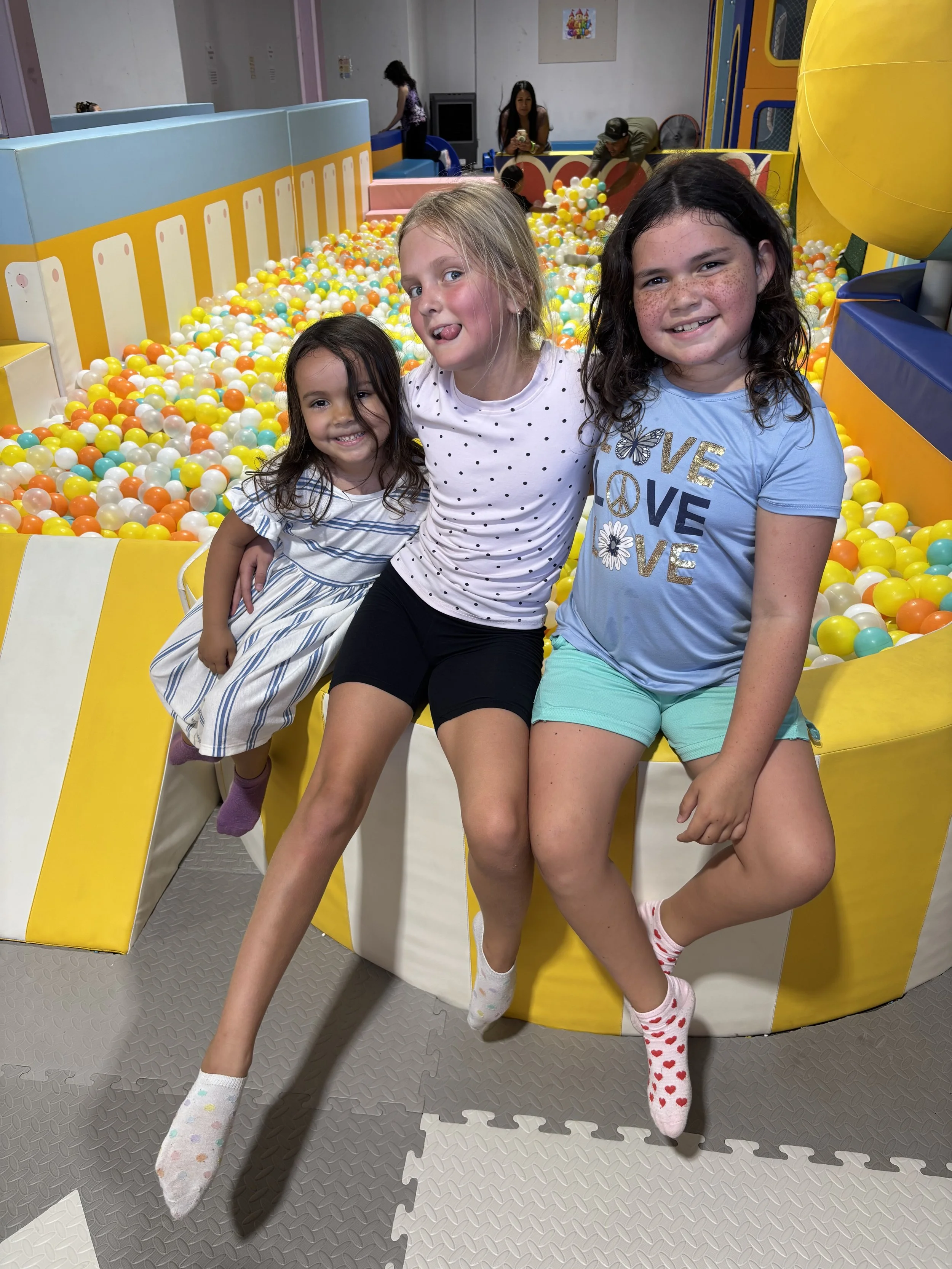 Three young girls sitting inside a ball pit at an indoor play area, smiling and having fun.