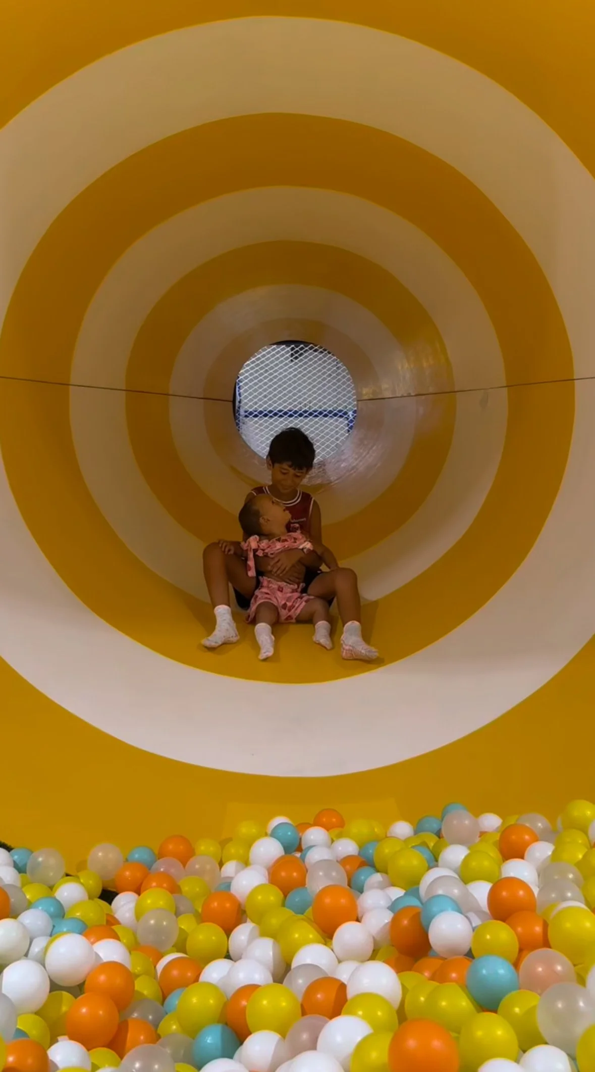 Children playing inside a yellow and white striped tunnel at a ball pit, with colorful plastic balls at the entrance.
