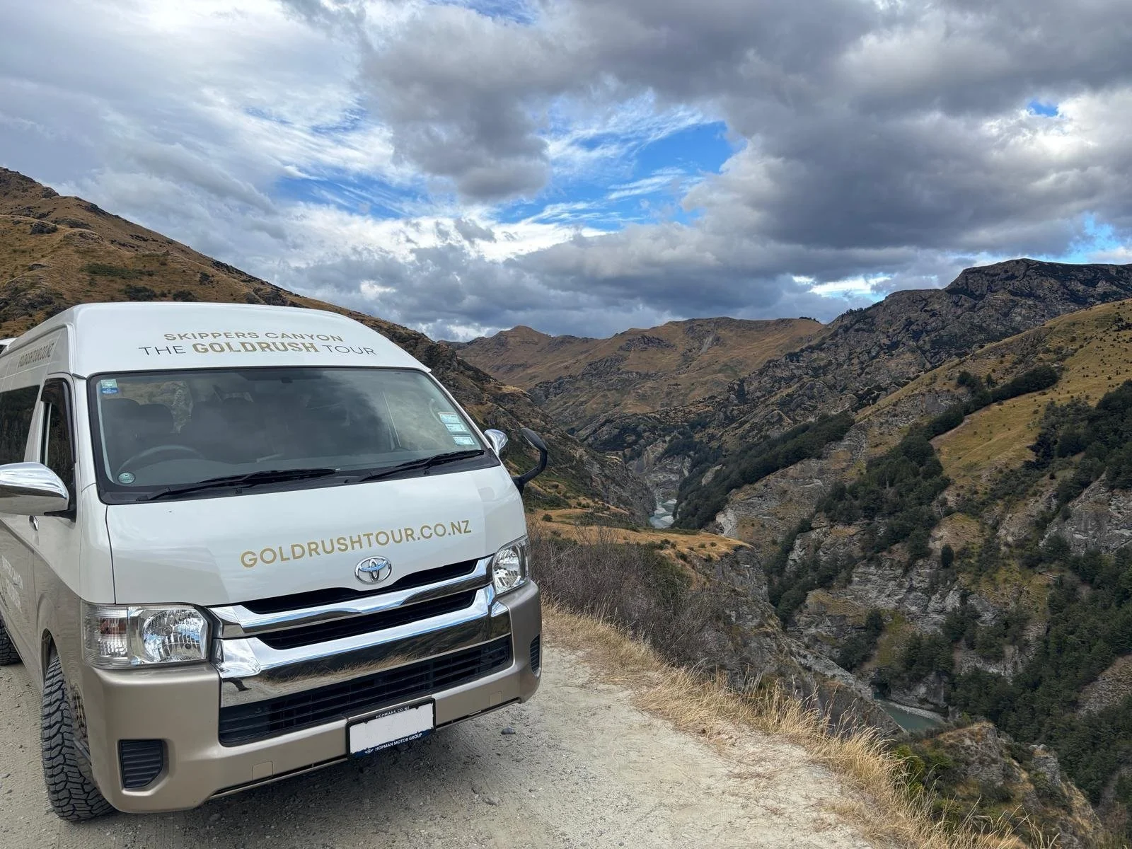 A silver Toyota van with the words "Skippers Canyon The Goldrush Tour" and "goldrushtour.co.nz" on the front, parked on a dirt road overlooking a deep canyon with rugged hills and a river at the bottom, under a cloudy sky.
