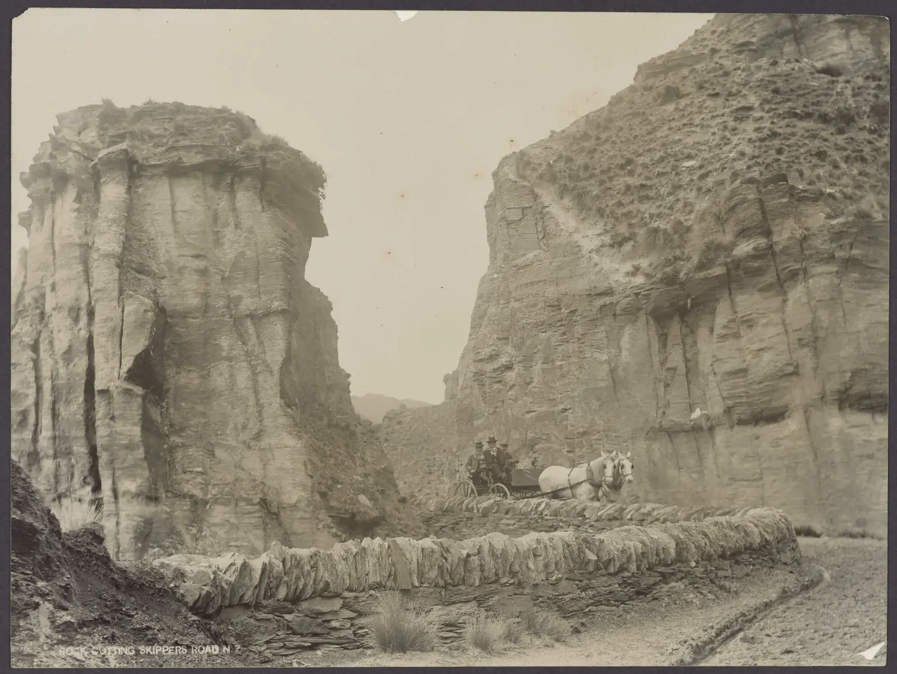 Old black and white photograph of a canyon with steep rock walls on either side and a dirt road running through the middle. A horse-drawn carriage with people is traveling on the road, and there's a small creek or stream at the bottom of the canyon, with a stone-lined bank.