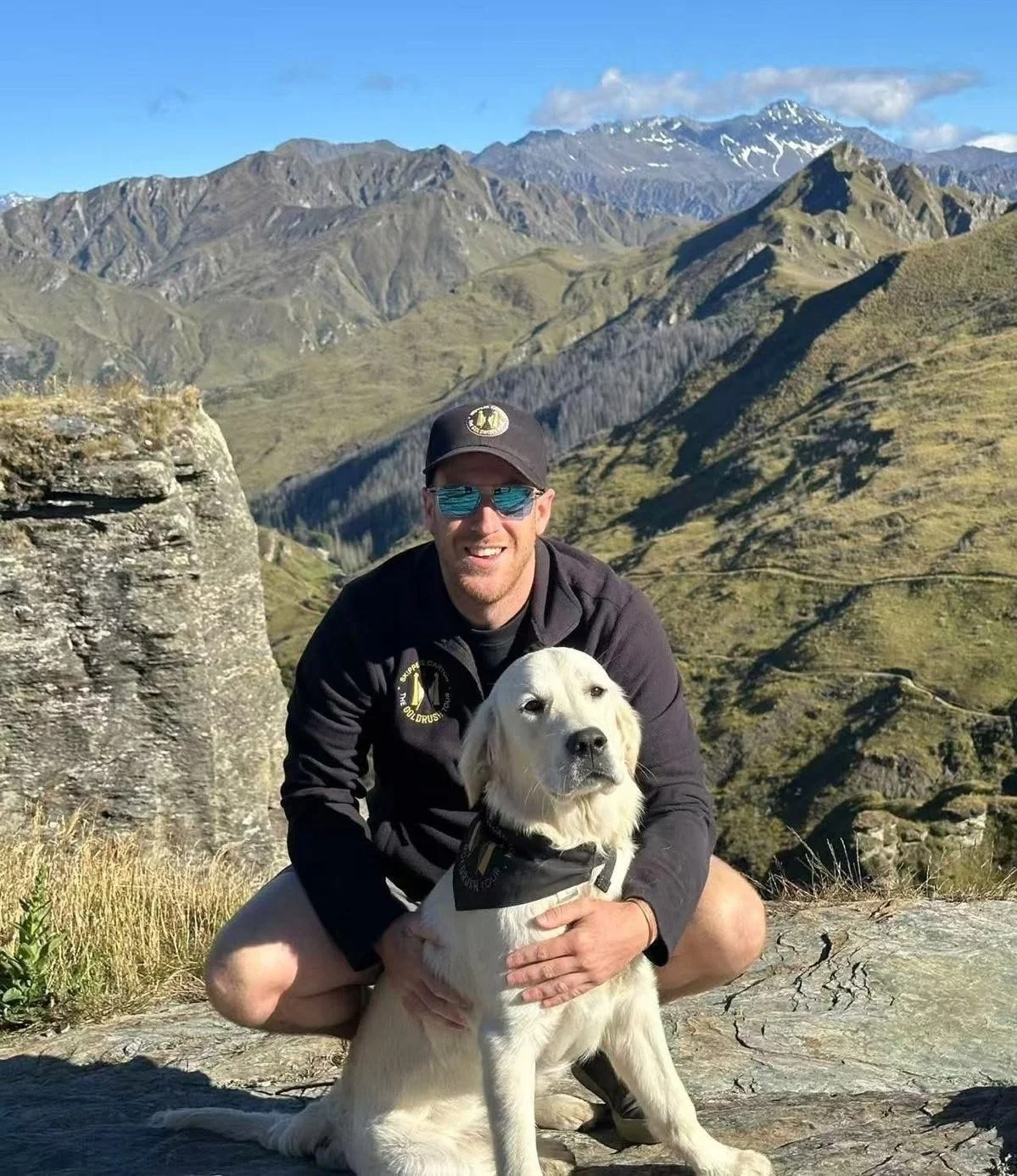 A man with sunglasses and a cap smiling while sitting with a golden retriever in front of a mountainous landscape with green hills and snow-capped peaks.