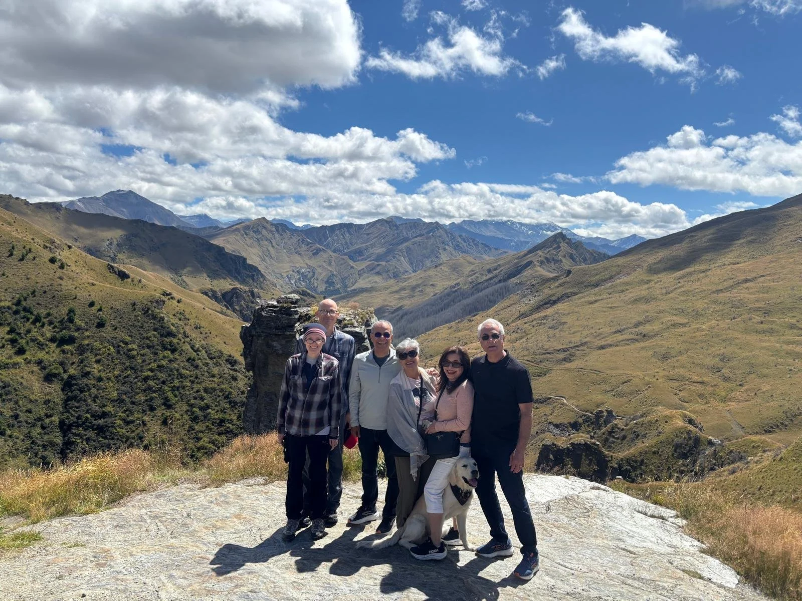 Group of six people with a dog standing on a rocky area in a mountainous landscape with hills and mountains in the background under a partly cloudy sky.