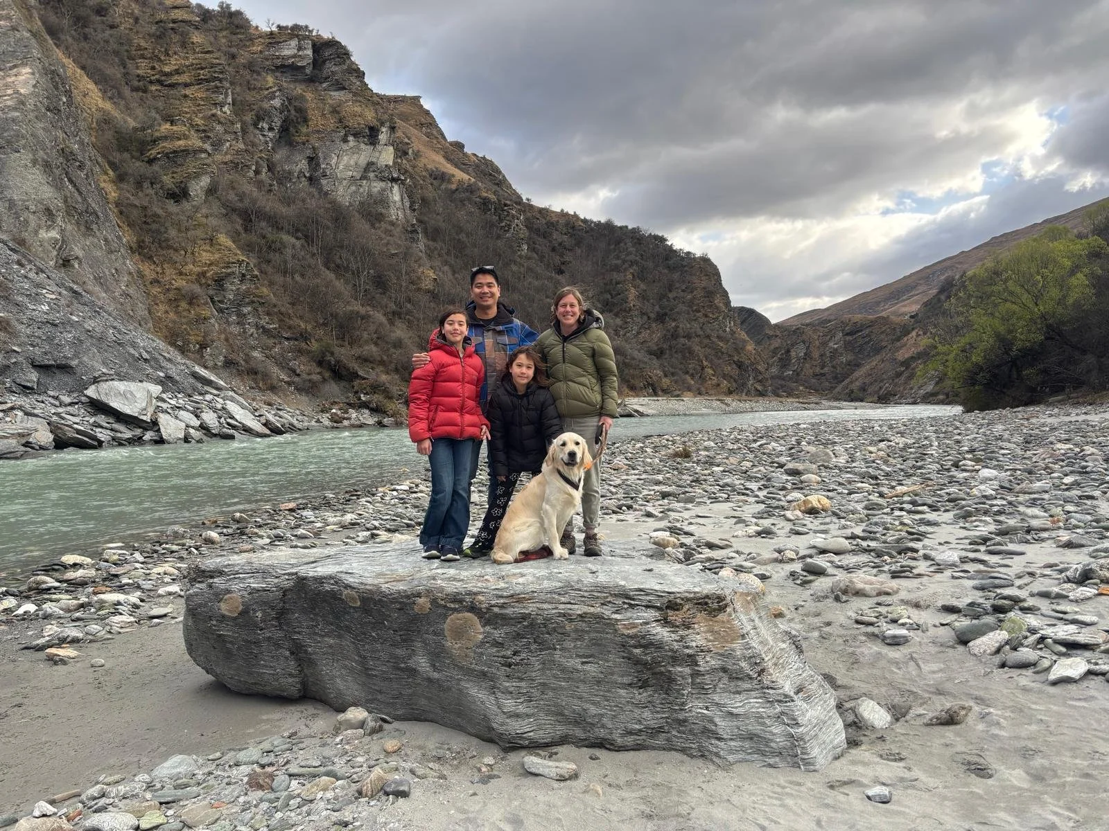 A family of four with a dog posing on a large rock by a river in a mountainous landscape with overcast sky.