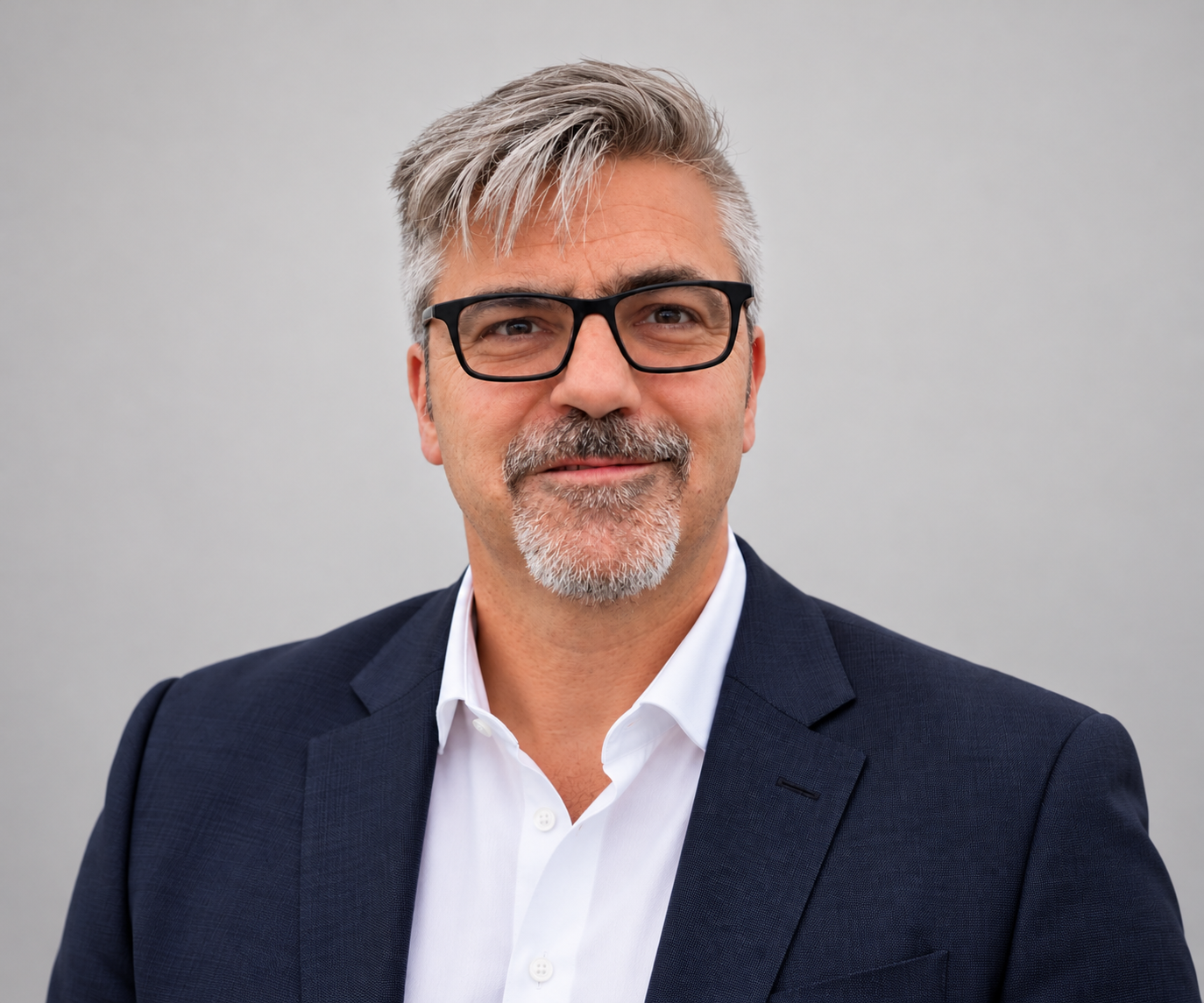 A middle-aged man with gray hair and a beard wearing black glasses, a dark blue suit, and a white shirt, smiling against a plain gray background.