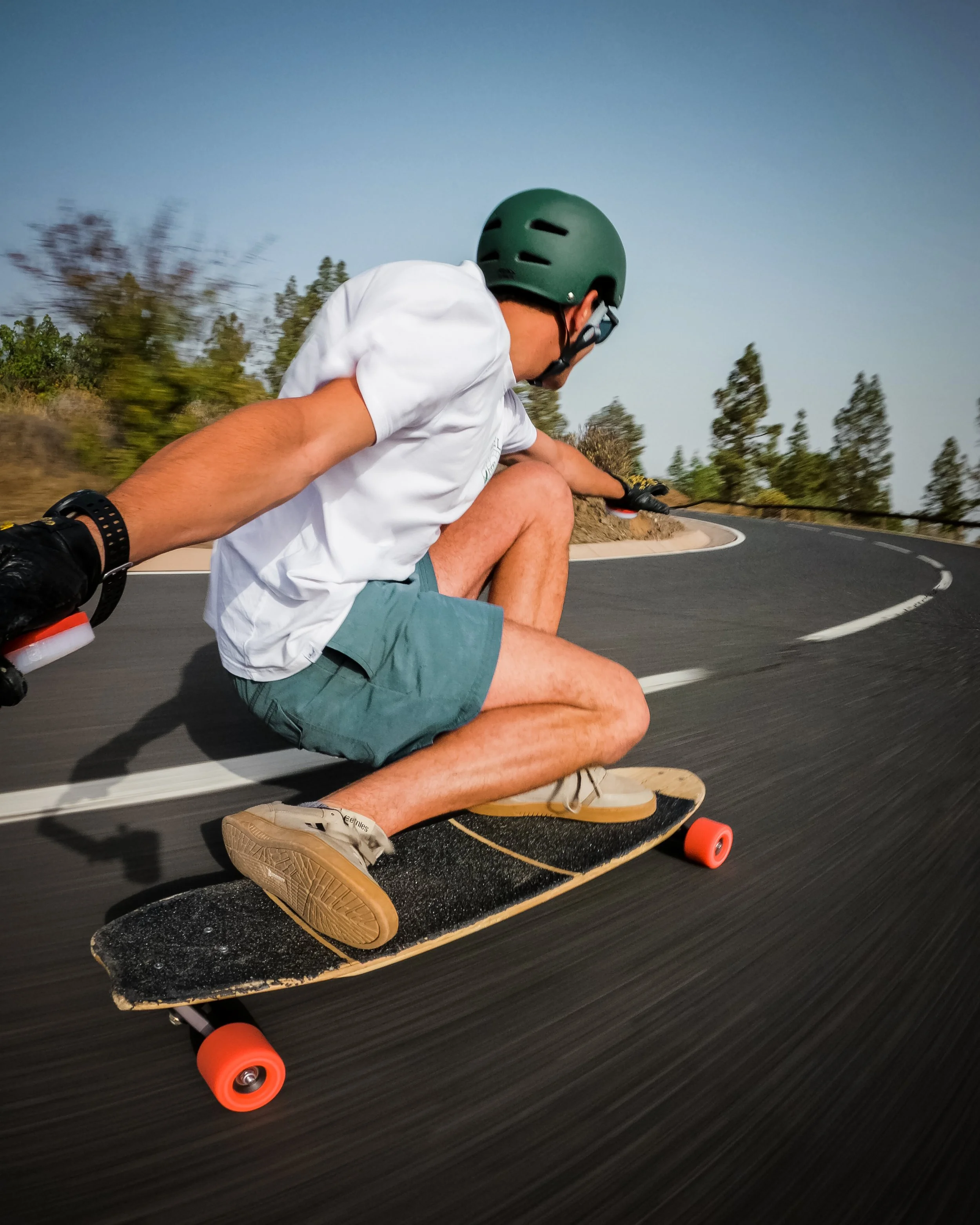Skateboarder wearing a green Elite 2.0 helmet, white T-shirt, and teal shorts, riding a skateboard on a curved road, with trees and a clear sky in the background.