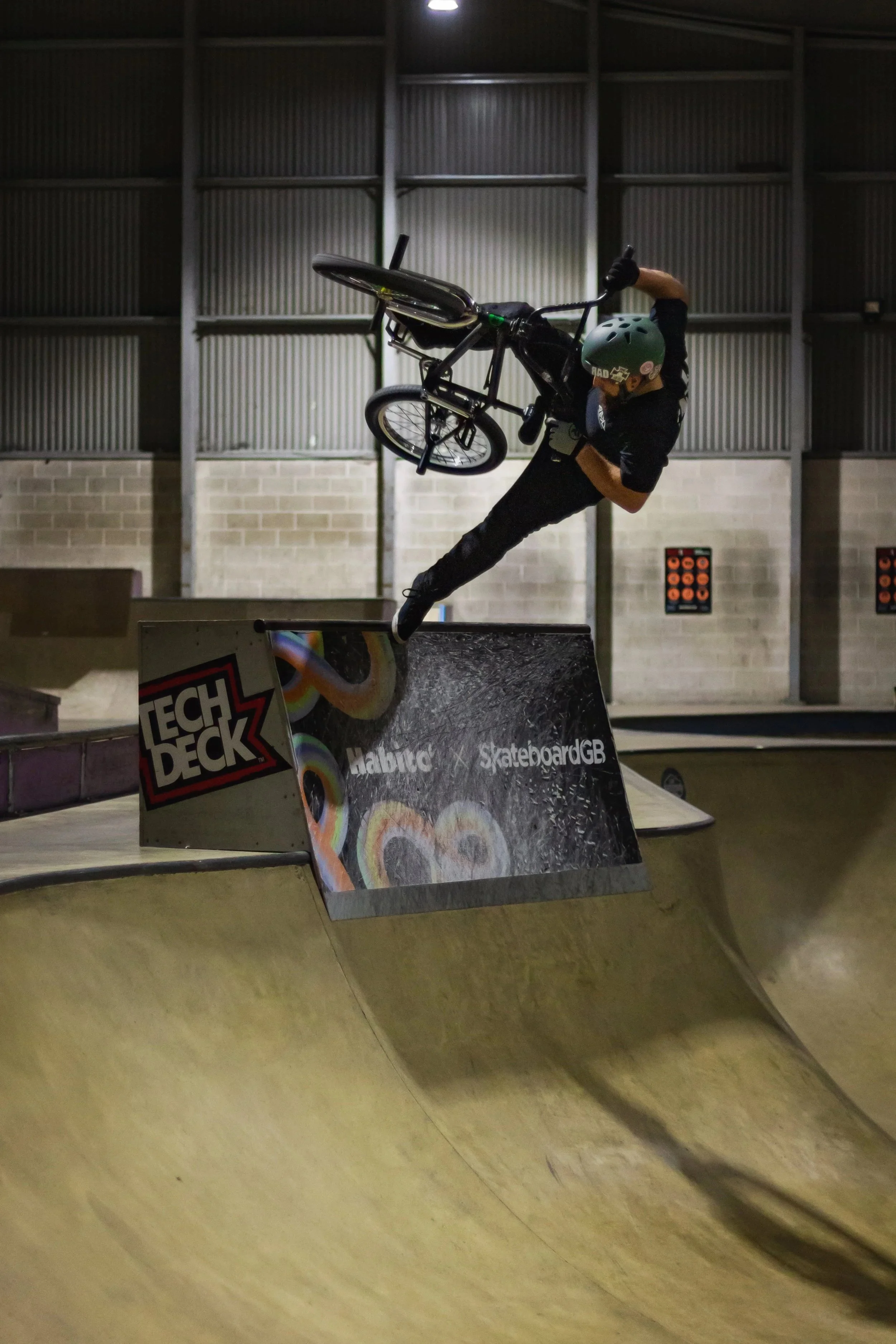 A person in a black shirt and REKD helmet is performing a BMX stunt while airborne at an indoor skate park.