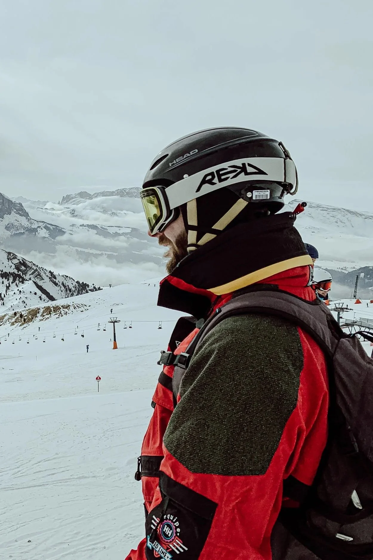 A person in winter gear, wearing a helmet and goggles, standing on a snowy mountain slope with ski lifts and mountain peaks in the background.